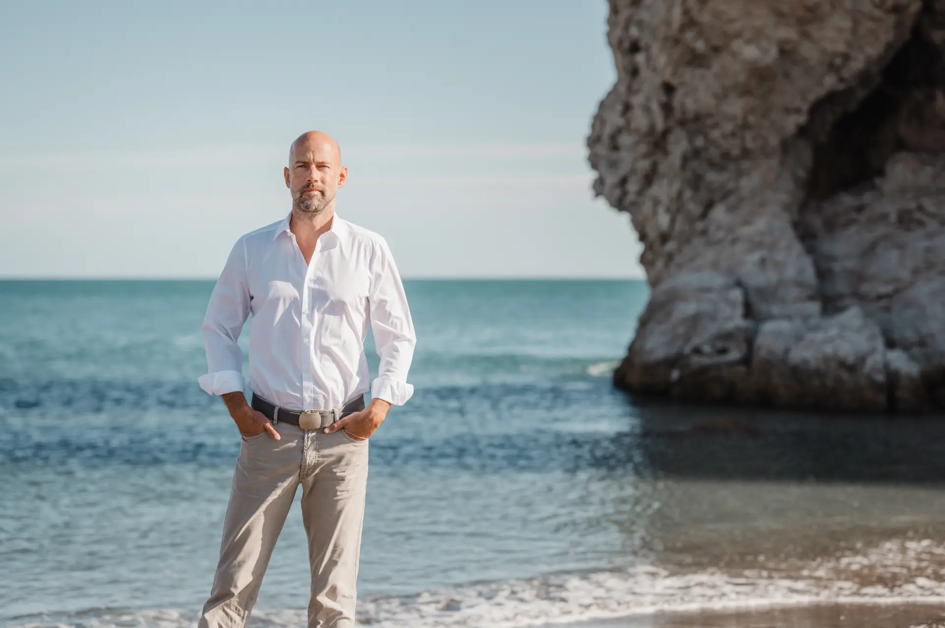 Man standing at the beach in a white shirt and beige pants, with the sea and a rocky cliff to the side behind him.